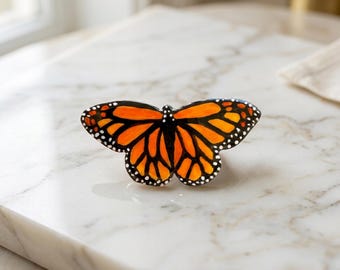 May include: A decorative butterfly with orange and black wings, detailed with white spots. The butterfly is set against a white marble surface, showcasing its vibrant colors and intricate design. The butterfly's wings are spread, giving the impression of flight.