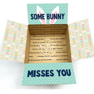 May include: A light blue and white patterned cardboard box with bunny ears and the text "Some Bunny Misses You". The box is open and the inside is lined with a light brown paper with a pattern of colorful Easter eggs.