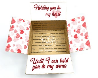 May include: A cardboard box with a heart pattern on the sides. The box is open and the inside has a message that reads "Holding you in my heart Until I can hold you in my arms".