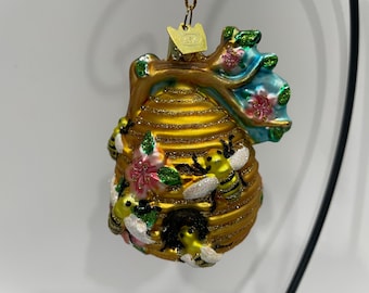 May include: A decorative ornament shaped like a golden beehive, adorned with bees, flowers, and a blue and green floral accent. The ornament is suspended by a gold chain and features a small tag.
