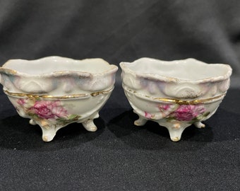 May include: Two white porcelain bowls with gold trim and pink rose floral designs. The bowls have three legs and a scalloped rim.