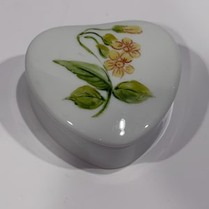 May include: A heart-shaped, white ceramic trinket box with a lid. The lid is decorated with a floral design featuring yellow flowers and green leaves. The box is likely used for storing small jewelry items or keepsakes.