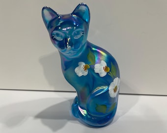 Peut inclure: Figurine de chat bleu irisé ornée de fleurs blanches et de feuilles vertes. Le chat est assis, les oreilles dressées. La figurine a une finition brillante et est probablement en verre ou en céramique.
