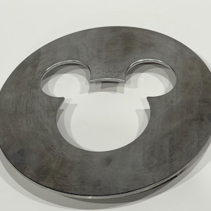 May include: A circular, metallic gray object with a cut-out design of Mickey Mouse's head and ears. The object is made of a solid, possibly metal, material and has a brushed texture. The design is centered within the circle.