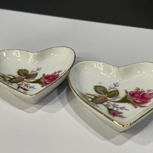 May include: Two heart-shaped, white porcelain dishes with a gold trim. Each dish features a floral design with pink roses, green leaves, and purple accents. The dishes are likely for holding small items.