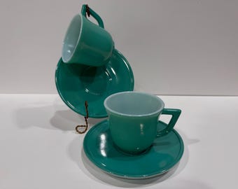 May include: Two teal glass cups and matching saucers. One cup rests on a saucer, while the other is positioned above a second saucer, held by a thin brown cord. The cups feature angular handles and a smooth, glossy finish throughout.