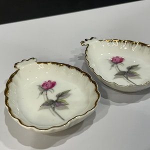 May include: Two white porcelain leaf-shaped dishes with gold trim. Each dish features a pink rose with green leaves. The dishes are likely for serving small portions of food or as decorative items.