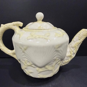 May include: Cream-colored ceramic teapot with a decorative design. The teapot features a curved handle with a dragon-like design, a spout, and a lid with a small knob. The teapot has a textured surface with raised floral patterns.