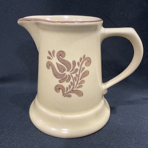 May include: A cream-colored ceramic pitcher with a brown floral design. The pitcher has a rounded body and a handle.
