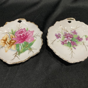 May include: Two small white porcelain leaf-shaped dishes with gold trim. One dish features a pink and yellow floral design, while the other has a purple floral design.