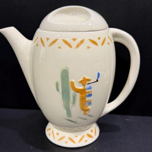 May include: Cream-colored ceramic teapot with a lid and handle. The teapot features a raised design of a cactus and a figure in a hat. Orange and blue accents decorate the teapot, including a pattern around the top and base. The spout is on the left side.