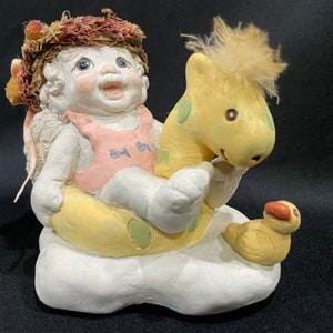 May include: A small, white ceramic figurine of an angel wearing a pink swimsuit and riding a yellow giraffe on a yellow inflatable ring. The angel has a halo and brown hair. The giraffe has green spots. The figurine is sitting on a white cloud.
