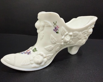 May include: A white ceramic shoe-shaped vase with embossed floral detailing. The vase features a high heel and is adorned with painted purple and green floral accents. The design includes roses and leaves, creating a vintage aesthetic.