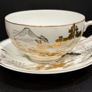 May include: White teacup and saucer set with gold landscape design. The cup and saucer feature a mountain and tree scene. The rim of the cup and saucer are trimmed in gold. The handle of the cup is white.