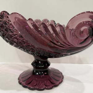 May include: A deep purple glass bowl with a pedestal base. The bowl features a scalloped rim, a swirling scroll design on the sides, and a textured diamond pattern on the exterior. The base is circular with a radiating petal-like pattern.