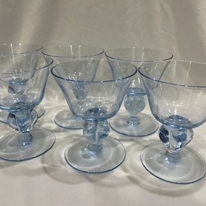 May include: Six light blue glass goblets with a rounded bowl and a stemmed base. The goblets have a unique design with a bulbous knob at the base of the stem.