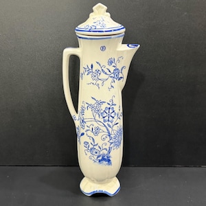 May include: A tall, cream-colored ceramic pitcher with a matching lid, featuring a blue floral pattern. The vessel has a slender, cylindrical body, a single side handle, and a flared spout. The blue design depicts delicate flowers and vines.