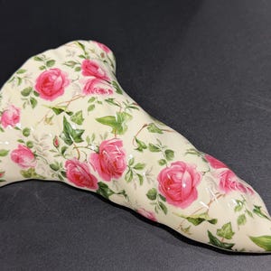 May include: A ceramic vase with a cream-colored base, adorned with a pattern of pink roses and green leaves. The vase has a unique, elongated shape, and the floral design covers the entire surface.