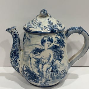 May include: A ceramic teapot with a lid, painted in blue and white. The teapot features a detailed cherub design on the body and floral patterns on the lid and spout. The handle is curved and decorated with blue accents.