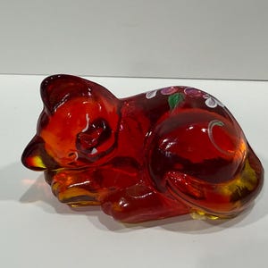 May include: A red glass cat figurine in a curled-up sleeping position. The figurine has a glossy finish and is decorated with painted flowers on its back. The cat's form is detailed, with visible ears, nose, and paws.