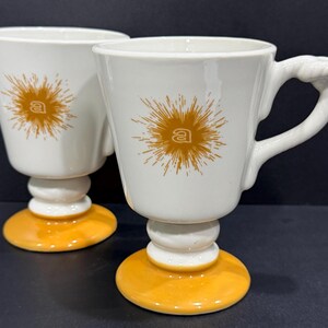 May include: Two white ceramic mugs with a pedestal base and a yellow circular foot. Each mug features a gold-colored sunburst graphic with a lowercase letter a in the center. The handle of each mug has a textured, rope-like design detail.