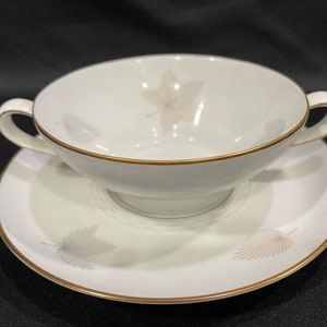 May include: A white porcelain soup bowl with a gold rim and a delicate leaf design. The bowl is sitting on a matching saucer with a gold rim and leaf design.