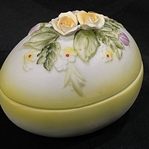 May include: A white ceramic egg-shaped trinket box with a lid. The lid is decorated with a floral design of yellow roses and green leaves.