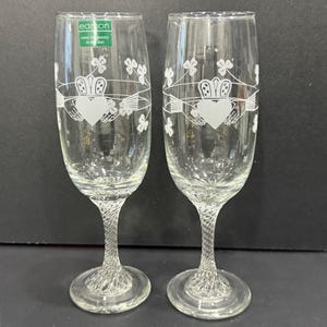 May include: Two clear champagne flutes with a white Claddagh design, featuring a heart, crown, and hands, along with shamrocks. The stems have a twisted design. The glasses have a label that says "Hand Designed in Ireland."