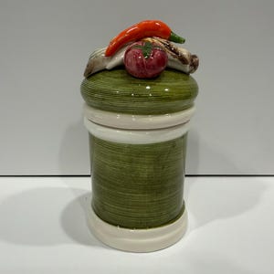May include: A ceramic jar with a green, textured body and a white base and rim. The lid is decorated with a red chili pepper, a red tomato, and other vegetables. The jar is designed to resemble a spool of thread.