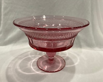 May include: A pink glass pedestal bowl with a diamond pattern. The bowl has a wide, shallow base and a tall, slender stem.