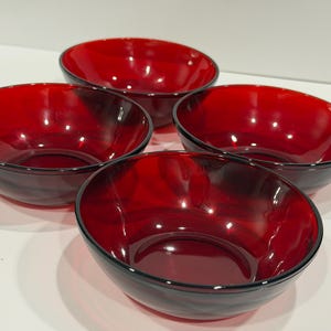 May include: Four red glass bowls with a glossy finish. The bowls have a deep red color and a black rim. The bowls are round and appear to be made of a thick glass material. The bowls are suitable for serving food.