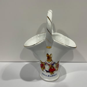 May include: White porcelain vase shaped like a basket with a gold handle. The vase features a colorful illustration of Mrs. Rabbit and her children from Beatrix Potter's children's book, "The Tale of Peter Rabbit".