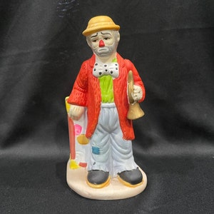 May include: A ceramic figurine of a sad clown holding a trumpet. The clown is wearing a red jacket, green vest, white pants, and a yellow hat. The figurine is standing on a white base with a colorful design.