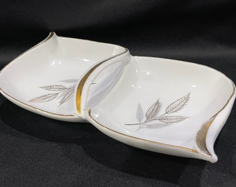 May include: A white ceramic serving dish with two sections and a gold handle. The dish has a silver leaf design.