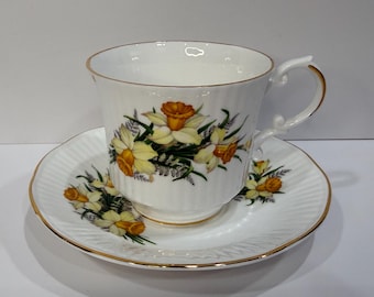 May include: White teacup and saucer set with a gold rim and a daffodil floral design. The teacup has a curved handle and the saucer has a ridged pattern. The daffodils are yellow and orange.