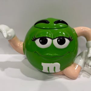 May include: A green ceramic teapot shaped like the M&M character with a glossy finish. The teapot features a round body, a white lowercase letter m, large eyes with black eyelashes, and light-colored arms with white gloves forming the spout and handle.