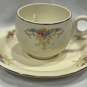 May include: A vintage teacup and saucer set with a floral pattern in shades of pink, yellow, and blue. The cup and saucer are both cream-colored with a gold trim.