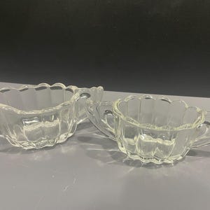 May include: A set of two clear glass creamer and sugar bowls with a scalloped edge and a fluted design.