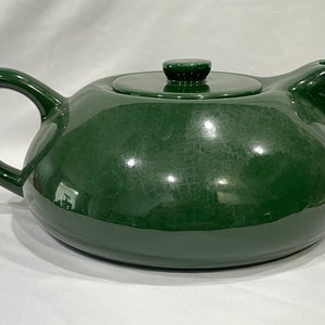 May include: A dark green ceramic teapot with a lid. The teapot has a rounded body and a spout on one side. The teapot is sitting on a white surface.