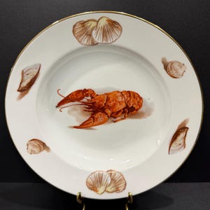 May include: White ceramic plate with a gold rim featuring a detailed illustration of a cooked lobster in the center. The plate is decorated with brown seashell illustrations around the rim, creating a nautical theme.
