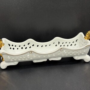 May include: A white porcelain serving dish with a pierced design and gold-colored handles. The dish has a scalloped edge and is supported by small feet. A gold trim accents the rim.
