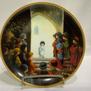 May include: A round porcelain plate with a gold rim features a painting of children gathered around a doorway. The children are dressed in colorful clothing and are looking up at a young boy standing in the doorway. The painting is set against a dark background with a light-colored doorway.