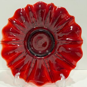 May include: A vibrant red glass plate with a ruffled, flower-like edge. The plate has a central, circular indentation and radiating lines. The glossy finish reflects light, highlighting the rich color and intricate design. A decorative piece for serving or display.