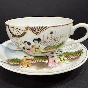 May include: White porcelain teacup and saucer set. The cup and saucer are decorated with a Japanese scene featuring figures in traditional attire, with gold accents. The cup has a handle and the saucer is round.