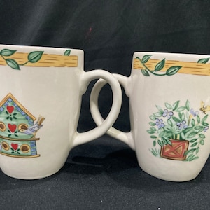 May include: Two white ceramic mugs with handles. The mug on the left has a green birdhouse with red hearts and blue birds. The mug on the right has a green plant with blue flowers in a red and white planter.