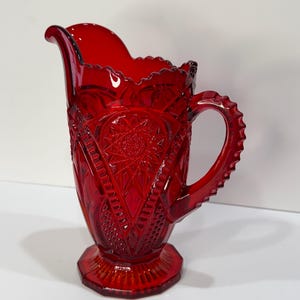 May include: A ruby red glass pitcher with a decorative, textured pattern. The pitcher has a scalloped rim, a detailed handle, and a hexagonal base. The design includes starburst and geometric shapes. A vintage piece.