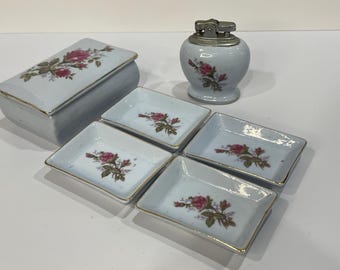 Può includere: Una collezione di oggetti in porcellana vintage, tra cui una scatola rettangolare con coperchio, quattro piccoli piatti rettangolari e un accendino rotondo. Ogni pezzo è azzurro con un motivo floreale di rose e rifiniture dorate.