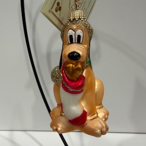 May include: A decorative Pluto Christmas ornament. The ornament features Pluto in a seated position, with a gold crown, a red and white outfit, and gold accents. The ornament is suspended by a gold string.