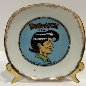 May include: A vintage Dogpatch U.S.A. ceramic plate with a gold rim, featuring a cartoon character on a light blue background. The plate is displayed on a gold-colored stand. The plate has the text "Dogpatch U.S.A." and "© 1968 CAPP ENTERPRISES, INC."