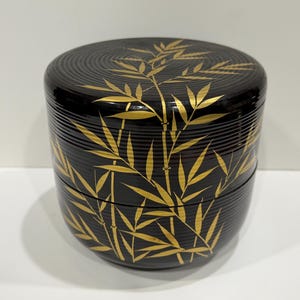 May include: A cylindrical black lacquer container with a lid, featuring a gold bamboo leaf and stalk pattern. The exterior surface has fine horizontal ridges throughout, and the design wraps continuously around the sides and the top of the lid.